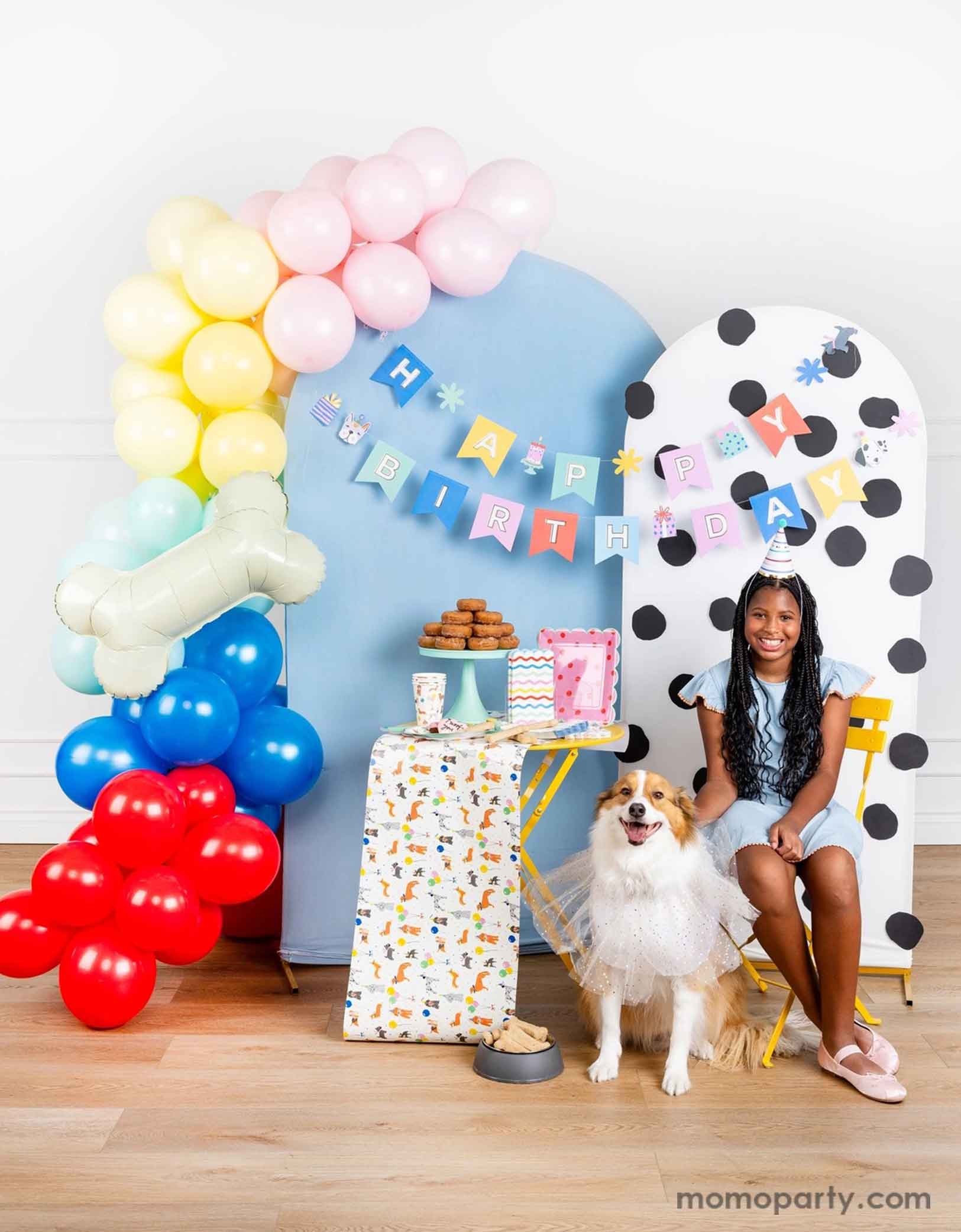 A dog themed party decoration set up featuring Momo Party's dog themed birthday supplies by My Mind's Eye. Including dog Happy Birthday party banner hung over a polka dotted and a pastel blue party backdrop with a colorful party balloon garland and a bone shaped foil balloon. On the table there are some dog themed party tablewares sugar cookies, treats and a donut tower. A girl in a birthday hat is sitting on the side with her dog happily celebrating her birthday.