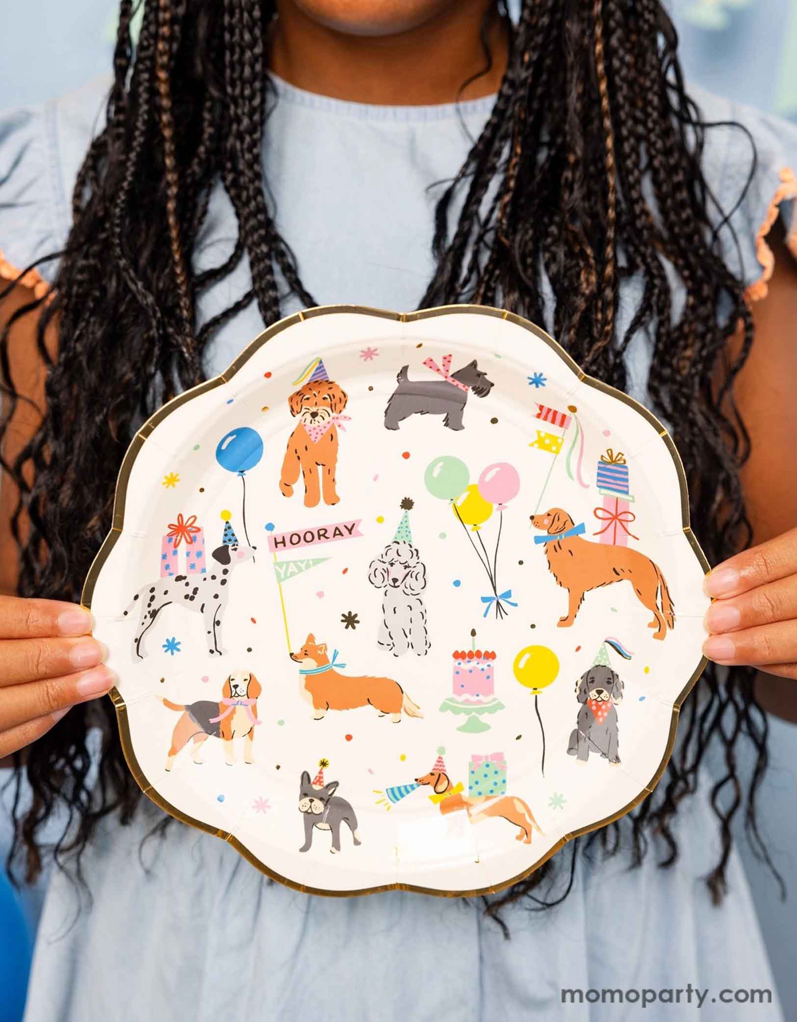 A girl in her pastel blue dress holding Momo Party's  9" Dog Party Plates by My Mind's Eye. Featuring adorable illustrations of different breeds of dogs with party hats, balloons and birthday cakes, these playful dog paper plates are perfect for any pet themed celebration.