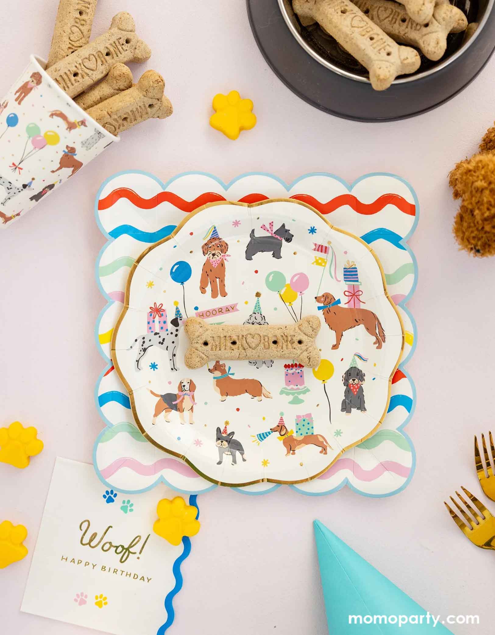 A festive and cheerful dog themed party table featuring Momo Party's dog party tableware collection including the dog paper plate paired with ric rac scalloped square plate, the dog party party cup, and woof napkins. With dog bone cookie treats around, this table is ready for a fun and playful birthday celebration for a pet lover!