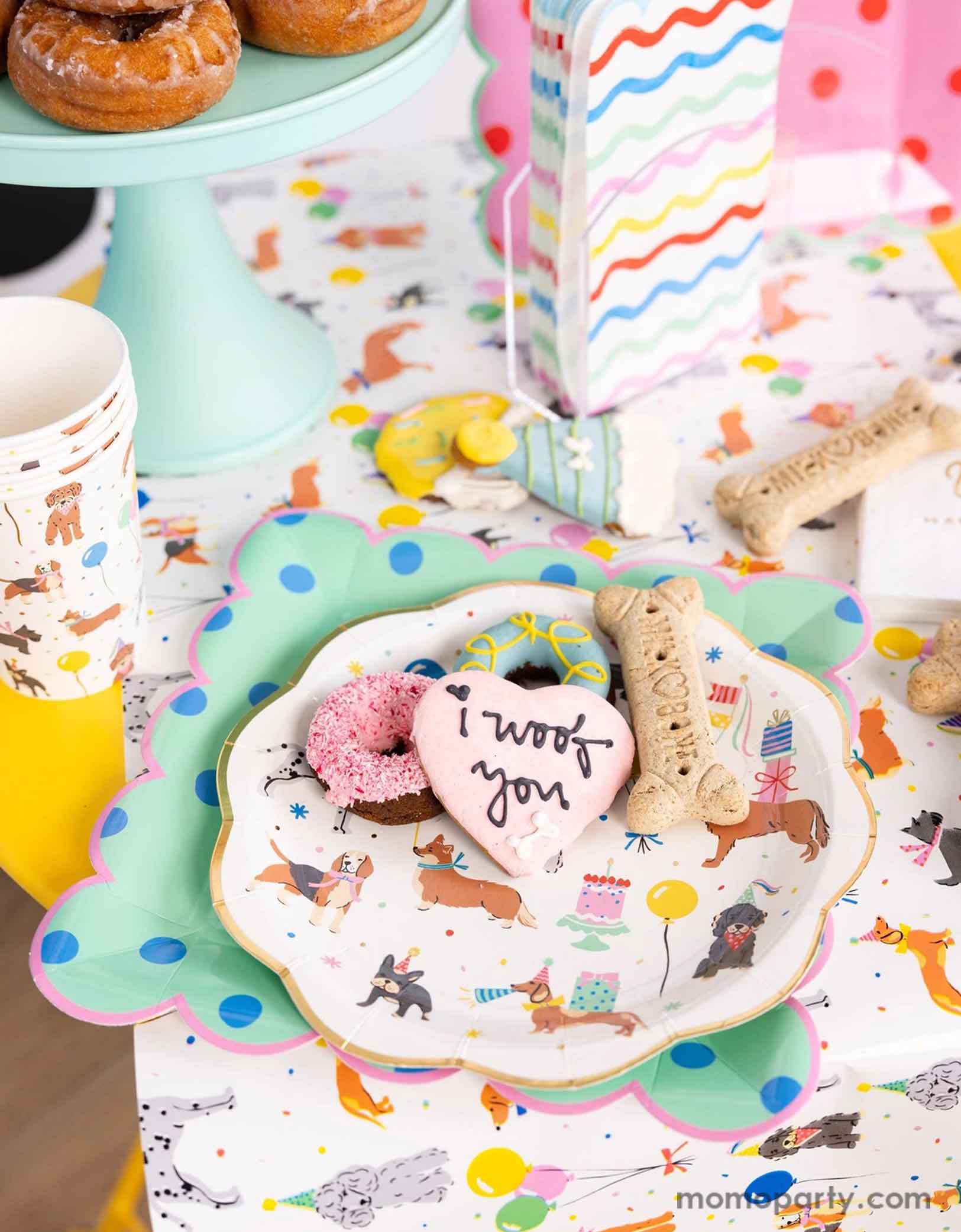 A festive and cheerful dog themed party table featuring Momo Party's dog party tableware collection including dog paper plates, party cups, napkins and table runner featuring adorable illustrations of different breeds of dogs with party hats, balloons and birthday cakes. With mint polka dot square plates and birthday festive sugar coated cookies that say "I Woof You" and donuts. This table is ready for a fun and playful birthday celebration for a pet lover!