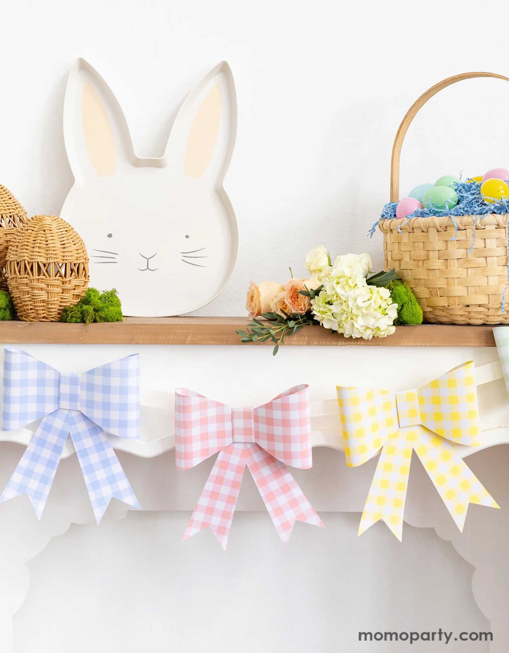 A mantel decorated with Easter decors including Momo Party's pastel gingham checkered bow party banner, Easter bunny shaped bamboo serving tray and some woven Easter egg decors, along with a basket filled with Easter eggs and spring floral bouquet, a simple yet adorable idea for spring home decor.