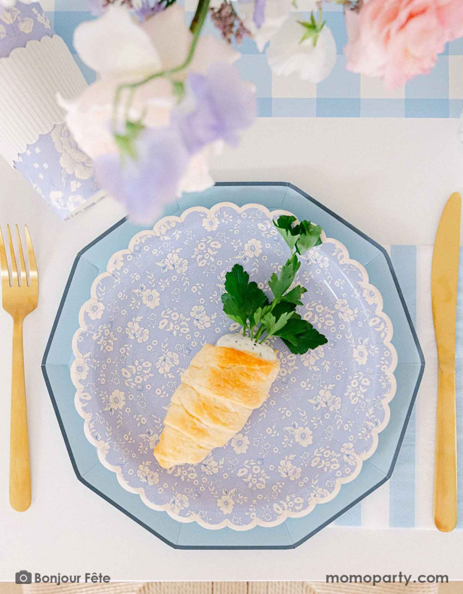 A beautiful Easter party tablescape featuring Momo Party's Easter themed party supplies in light blue including sky blue decagon dinner plates layered with blue ditsy floral scallop plate, with the matching paper cup, sky blue cabana striped guest napkins and gold foil utensils. On the table there's a food cup filled robin egg inspired candies on a blue gingham table runner. Along with the carrot inspired pastry, making this a perfect look for an elegant Easter party tableset. 