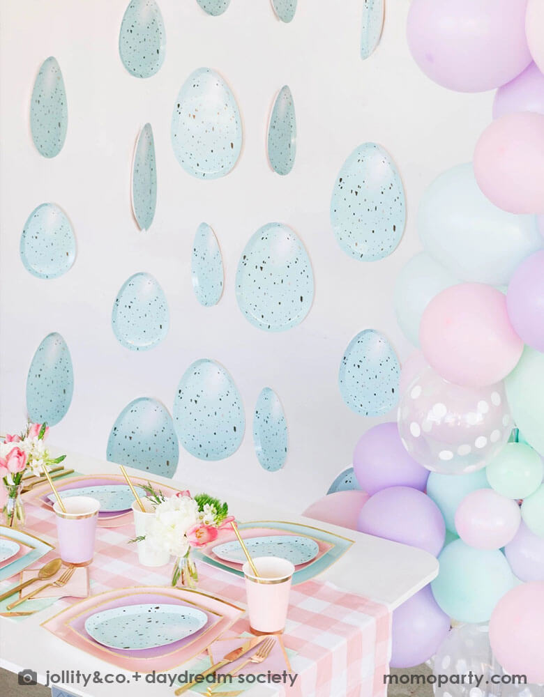 A spring inspired Easter party table decoration featuring Momo Party's pastel themed tableware on the party table including pastel mint & pink plates and robin egg shaped plates, lilac party cups, pink gingham table runner and gold utensils. Next to the table there's a pastel balloon garland next to some robin egg party garland hung on the wall, making this a great Easter party inspo for springtime.