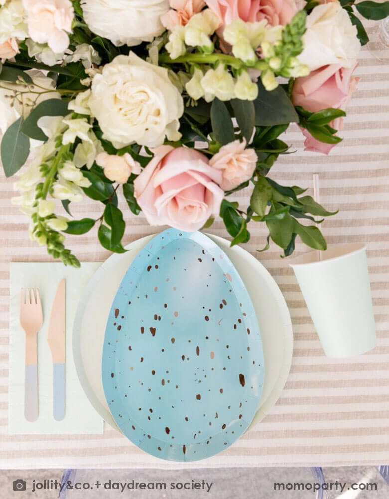 An closeup shot of Easter party table set up with Momo Party's Rockin' Robin Dinner Plates layered with pastel mint round plate, color matching paper cups, napkins, and pastel blue dipped wooden utensils, with a vase with flowers and on a light beige striped tablecloth for a modern chic Easter table placesetting.