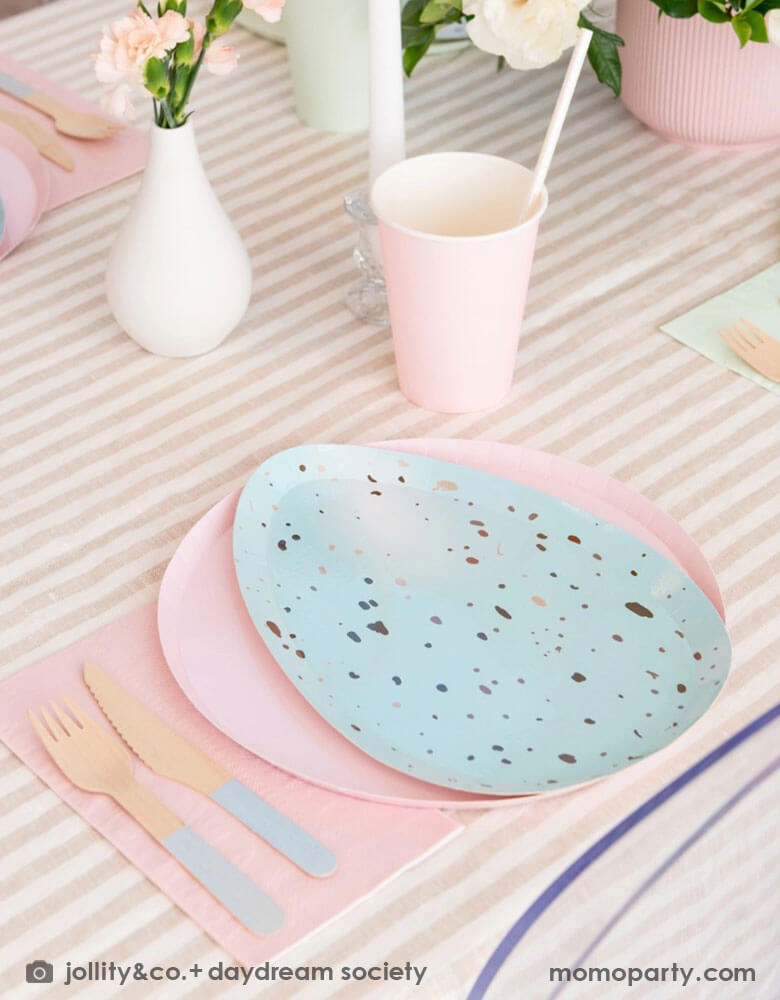 An closeup shot of Easter party table set up with Momo Party's Rockin' Robin Dinner Plates layered with pastel pink round plate, color matching paper cups, napkins, and pastel blue dipped wooden utensils, with vases with flowers and candles on a light beige striped tablecloth for a modern chic Easter table setting.