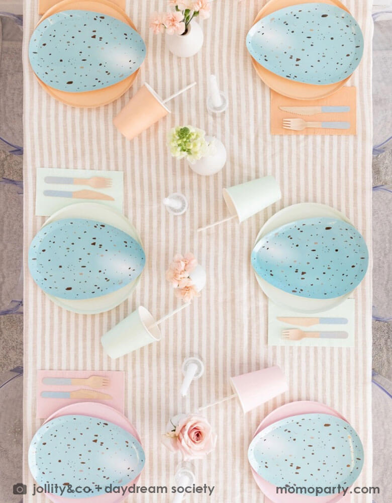 An overhead shot of Easter party table set up with Rockin' Robin Dinner Plates layered with pastel colored round plates in peach, mint and pink, with color matching paper cups, napkins, and pastel blue dipped wooden utensils, with vases with flowers and candles on a light beige striped tablecloth for a modern chic Easter table setting.