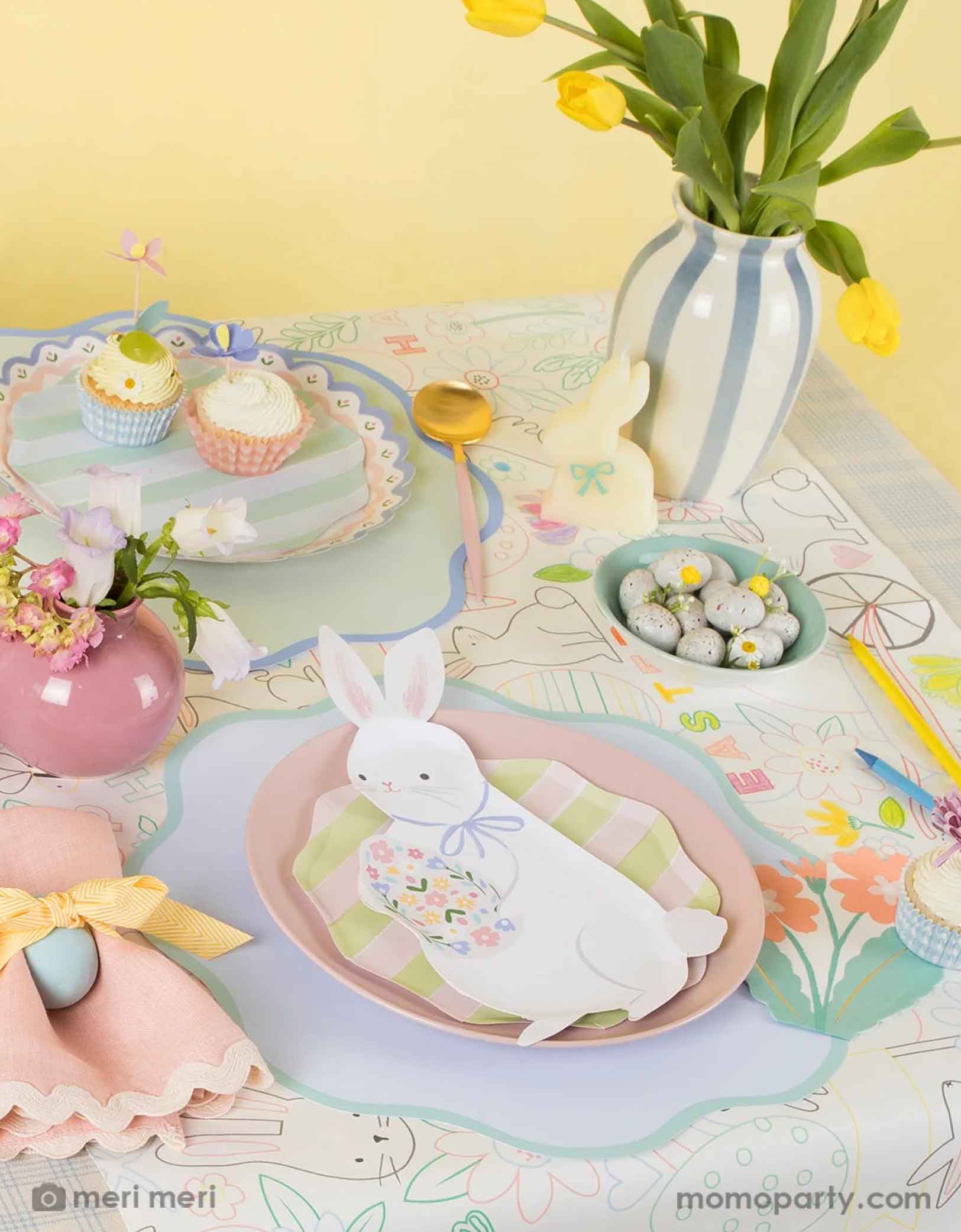 A kid's friendly Easter table setting featuring Momo Party's Easter party supplies by Meri Meri including the Easter bunny shaped plates, gingham checkered Easter cupcake kit, the Easter bunny coloring tablecloth, with pastel blue scalloped placemats, with spring tulip flowers and Easter egg shaped chocolate candies on the table, this makes a cute and simple placeset to celebrate Easter with your kids. 