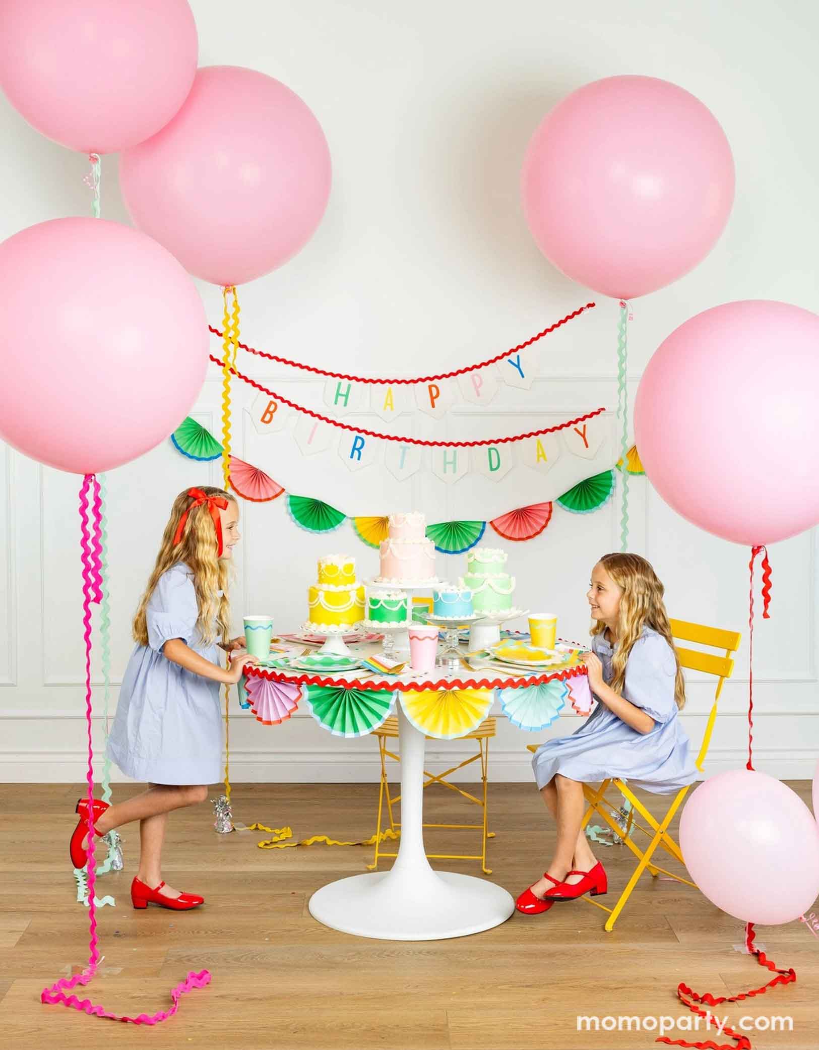 A cheerful and colorful birthday party set up featuring various festive birthday party supplies from Momo Party including rainbow colored tablewares of plates, cups, napkins on the table along with multiple pastel colored birthday cakes. The table is decorated with colorful bunting garland around and in the back of the wall hung a Happy Birthday felt garland and the colorful bunting banner. On the sides are some jumbo pink balloons floating above and two girls in baby blue dresses sitting at the table.