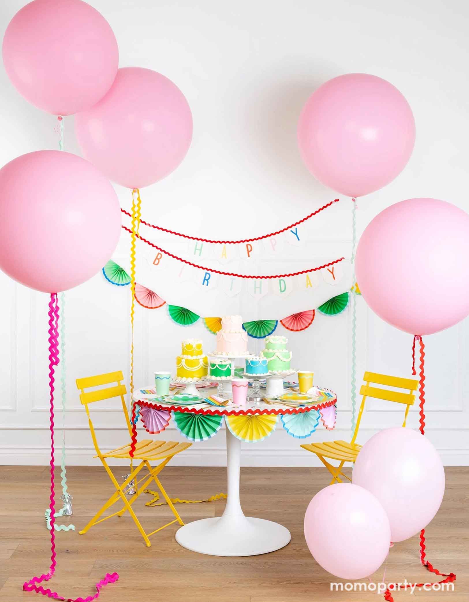 A cheerful and colorful birthday party set up featuring various festive birthday party supplies from Momo Party including rainbow colored tablewares of plates, cups, napkins on the table along with multiple pastel colored birthday cakes. The table is decorated with colorful bunting garland around and in the back of the wall hung a Happy Birthday felt garland and the colorful bunting banner. On the sides are some jumbo pink balloons floating above, making this a whimsical set up for kid's birthday bash!