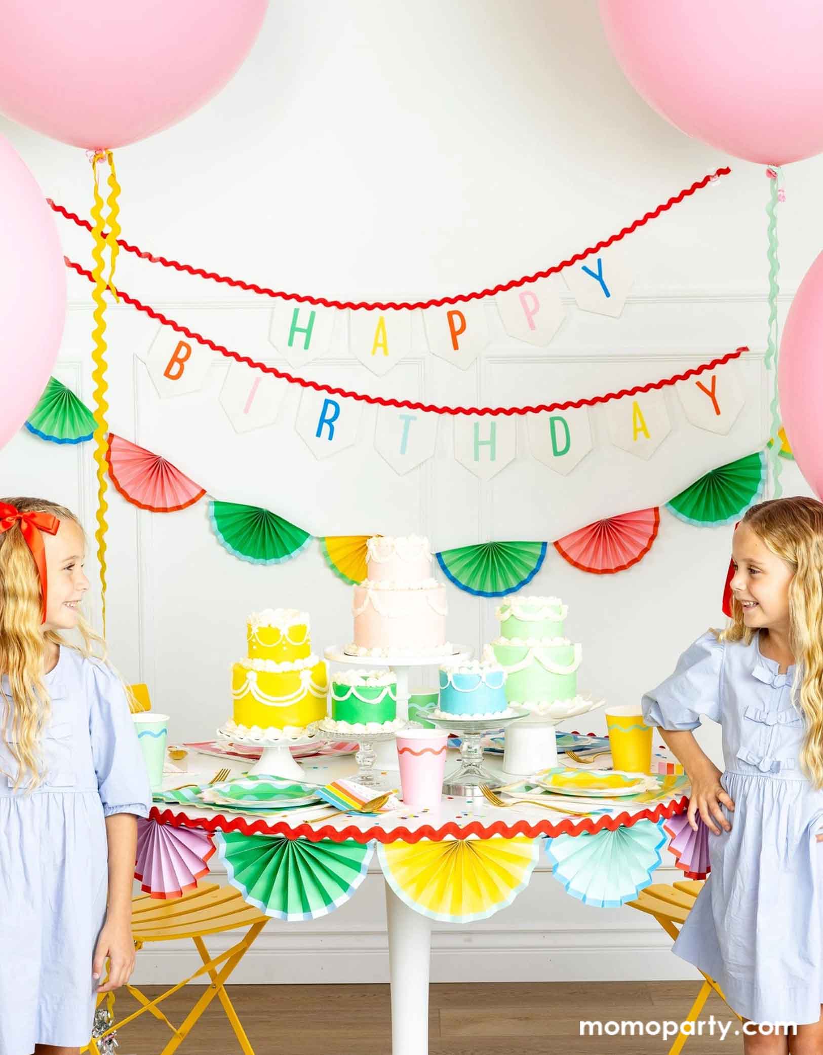 A cheerful and colorful birthday party set up featuring various festive birthday party supplies from Momo Party including rainbow colored tablewares of plates, cups, napkins on the table along with multiple pastel colored birthday cakes. The table is decorated with colorful bunting garland around and in the back of the wall hung a Happy Birthday felt garland and the colorful bunting banner. On the sides are some jumbo pink balloons floating above and two girls in baby blue dresses next to the table.