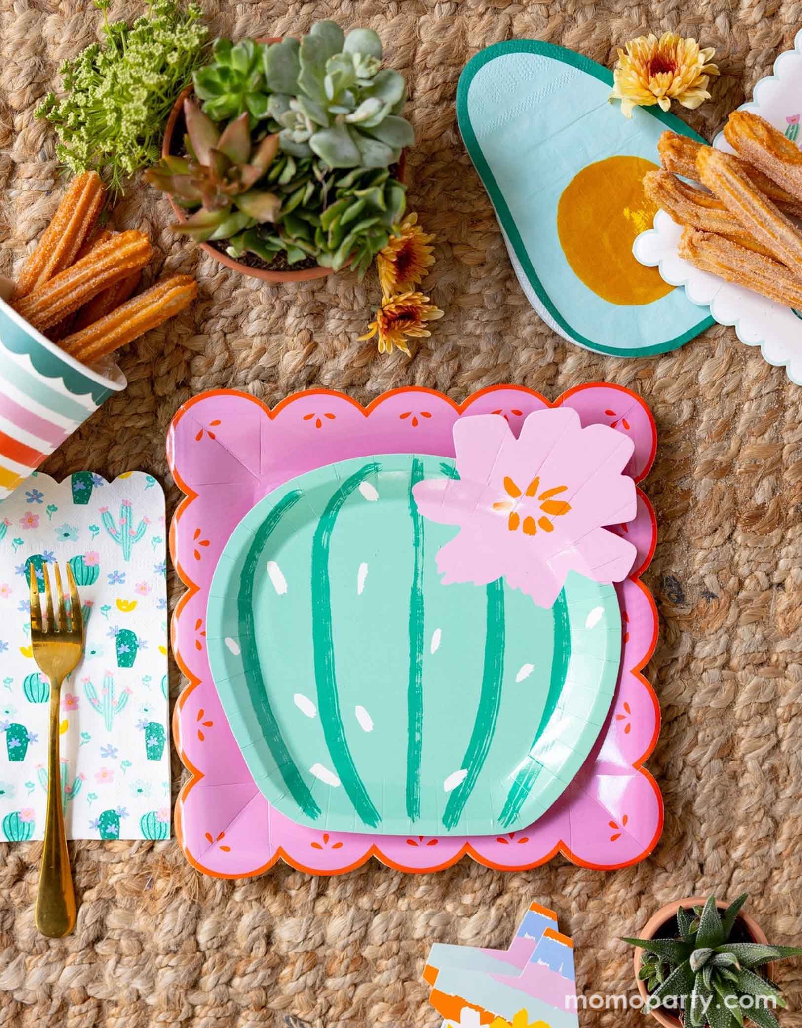 A overhead shot of a fun and playful fiesta themed party tablescape featuring Momo Party's fiesta themed party supplies including the pink Fiesta Papel paper plate, the cactus shaped plate, avocado shaped napkins, and the fiesta rainbow colored striped party cup filled with churros. On the table there are mini pots of cactus and succulent plants, making this a fun table setting idea for a playful yet modern fiesta themed celebration - perfect for girl's "first fiesta" themed first birthday party!