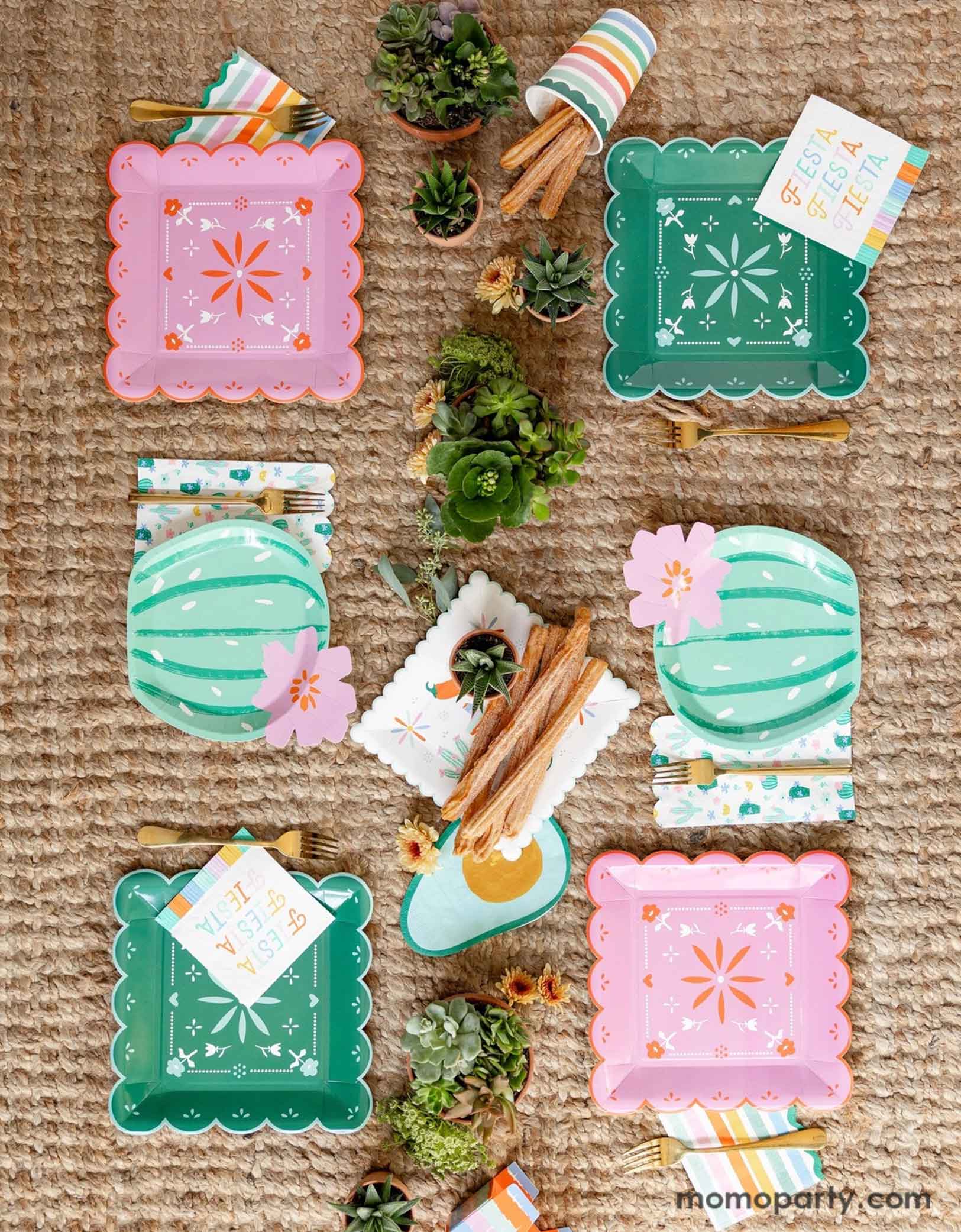 A overhead shot of a fun and playful fiesta themed party table featuring Momo Party's fiesta themed party supplies including the Fiesta Papel paper plates in mint and pink, the cactus shaped plates, fiesta fringe napkins, avocado shaped napkins and the fiesta rainbow colored striped party cup filled with churros. On the table there are mini pots of cactus and succulent plants, making this a fun table setting idea for a playful yet modern fiesta themed celebration!