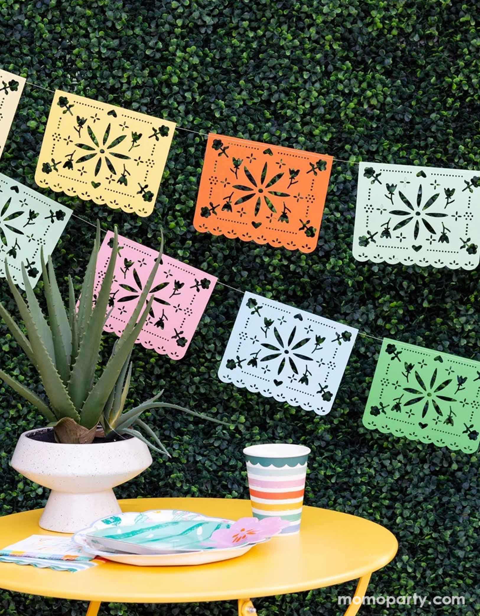 A festive Mexican themed celebration set up featuring Momo Party's Fiesta party supplies including the rainbow colored Fiesta Papel Picado Banner on the faux leaf wall, cactus shaped plate, fiesta fringe napkins and fiesta striped party cups on a bright yellow table. With a cactus/succulent plate as the centerpiece, this makes a simple yet modern look for a fun and playful fiesta celebration!