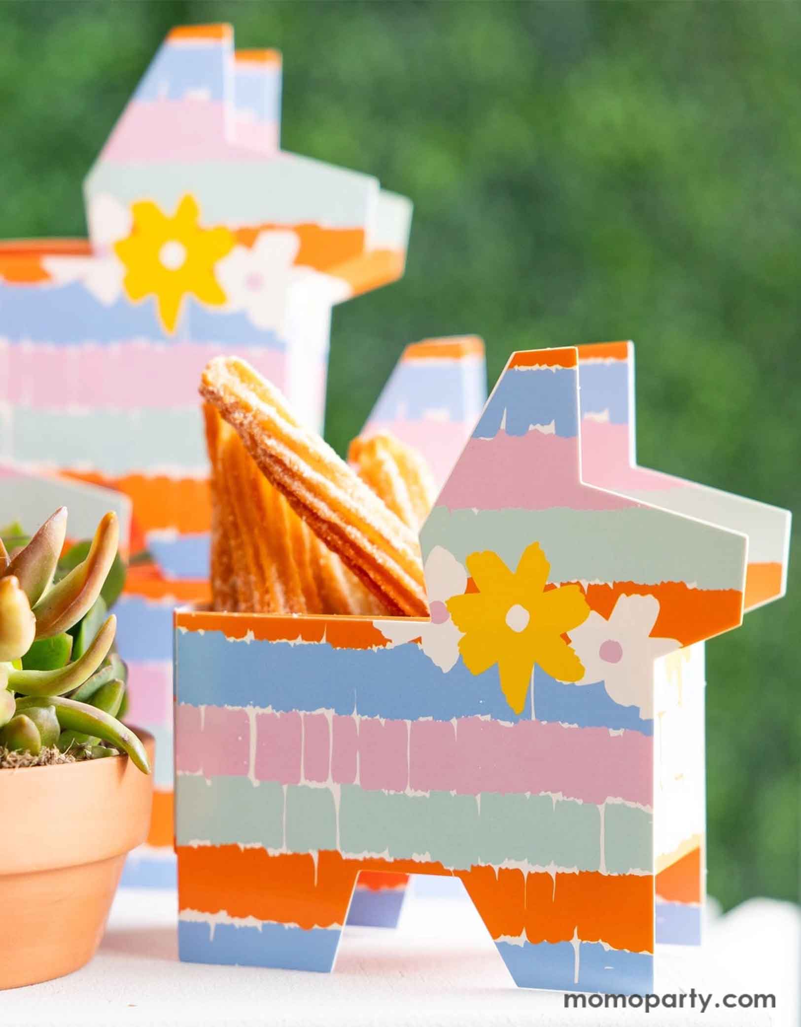 A festive Feista party table featuring Momo Party's fiesta pinata shaped treat boxes in colorful rainbow colors with fringe design. The treat boxes are filled with churros and are set next to a cactus plant, making them a perfect addition to a fun and playful kid's fiesta themed birthday celebration or a Cinco de Mayo party!