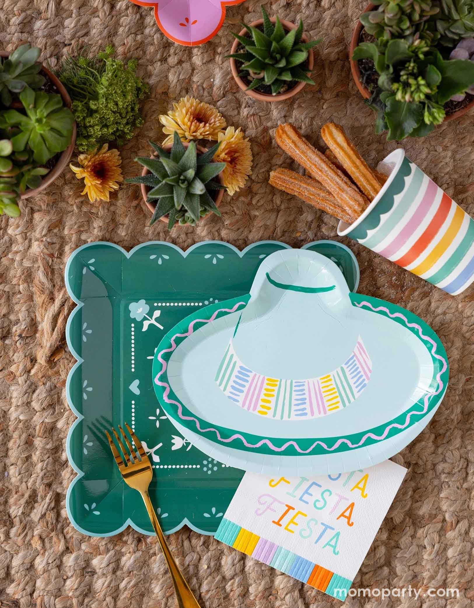 A overhead shot of a fun and playful fiesta themed party tablescape featuring Momo Party's fiesta themed party supplies including the Fiesta Papel paper plate, the sombrero hat shaped plate, fiesta fringe napkins, and the fiesta rainbow colored striped party cup filled with churros. On the table there are mini pots of cactus and succulent plants, making this a fun table setting idea for a playful yet modern fiesta themed celebration - perfect for kid's "first fiesta" themed first birthday party!