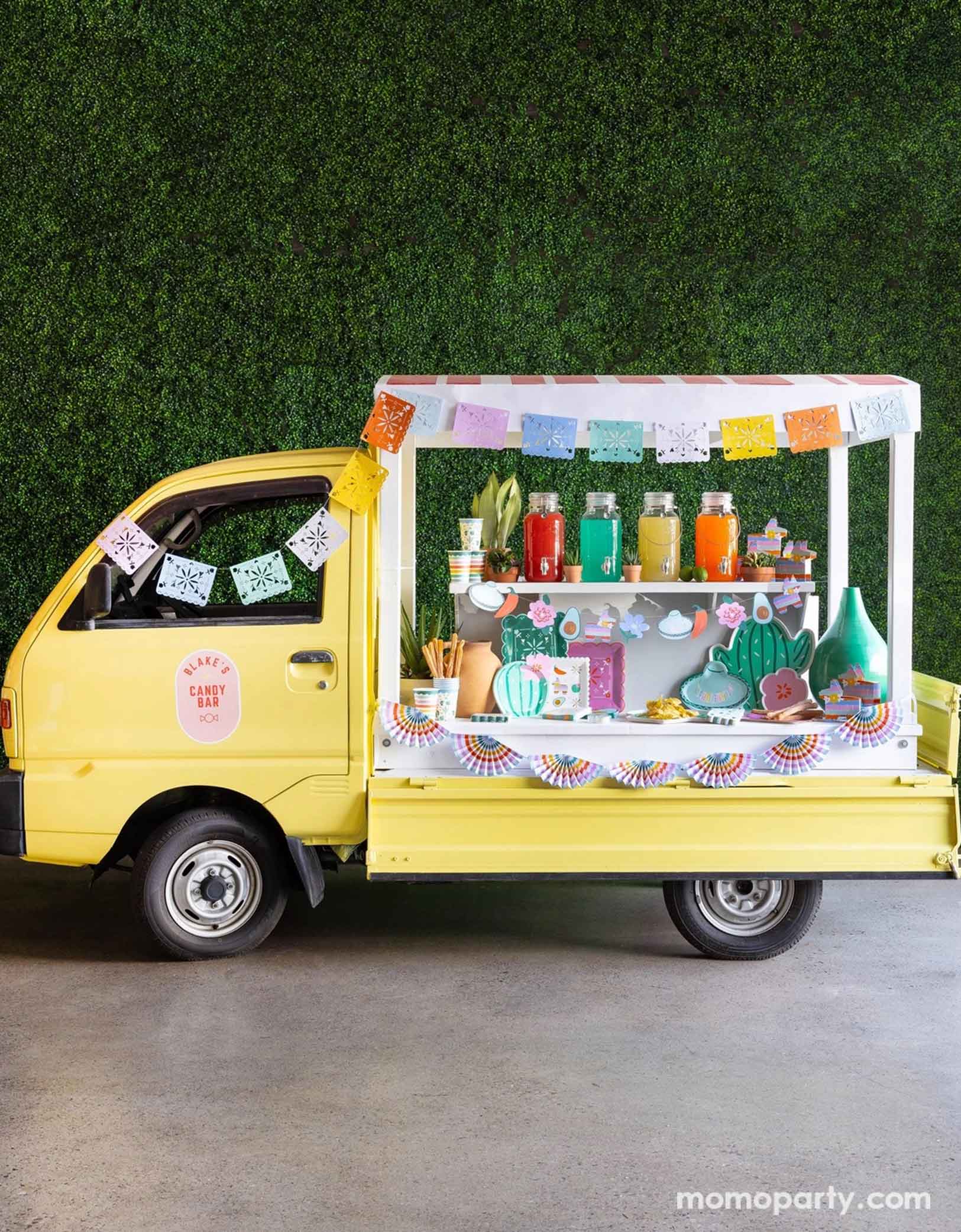 A cheerful yellow Mexican inspired truck is decorated with Momo Party's colorful fiesta party supplies including the Fiesta Papel Picado Banner, bunting banner hung across the truck. In the middle there a various fiesta party tablewares in display including cactus shaped plates, sombrero hat shaped plates, fiesta icon dinner plates, fringe napkins, striped party cups and pinata shaped treat boxes. On the truck there are also colorful drinks in glass pitchers ready to be served for the party guests.