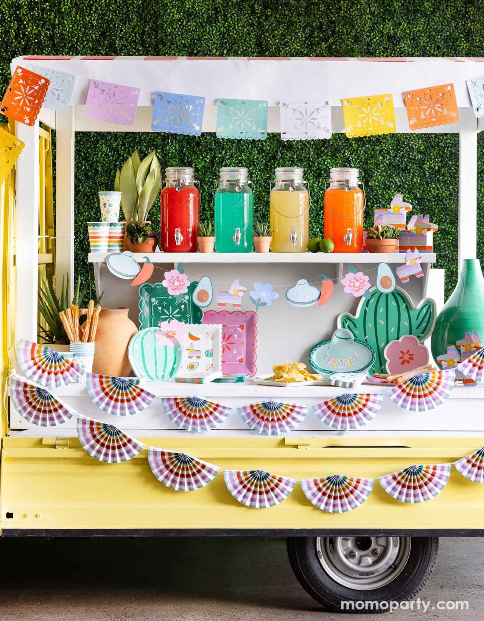 A cheerful yellow Mexican inspired truck is decorated with Momo Party's colorful fiesta party supplies including the Fiesta Papel Picado Banner, bunting banner hung across the truck. In the middle there a various fiesta party tablewares in display including cactus shaped plates, sombrero hat shaped plates, fiesta icon dinner plates, fringe napkins, striped party cups and pinata shaped treat boxes. On the truck there are also colorful drinks in glass pitchers ready to be served for the party guests.