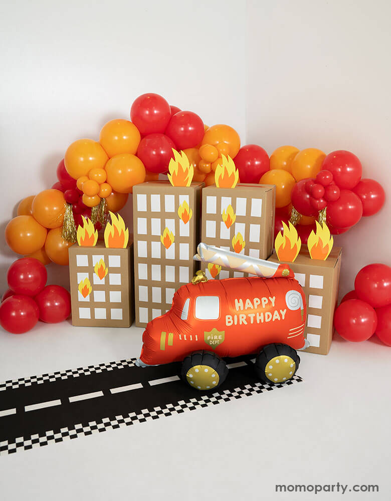 A fire truck themed birthday party balloon decoration featuring Momo Party's fire truck foil balloon and balloon garland in red and orange. Under the balloon garland there are playhouse buildings made out of packaging boxes with fire flame paper cutouts, making this a fun and playful decoration for kid's fire truck themed birthday party or classroom decoration!