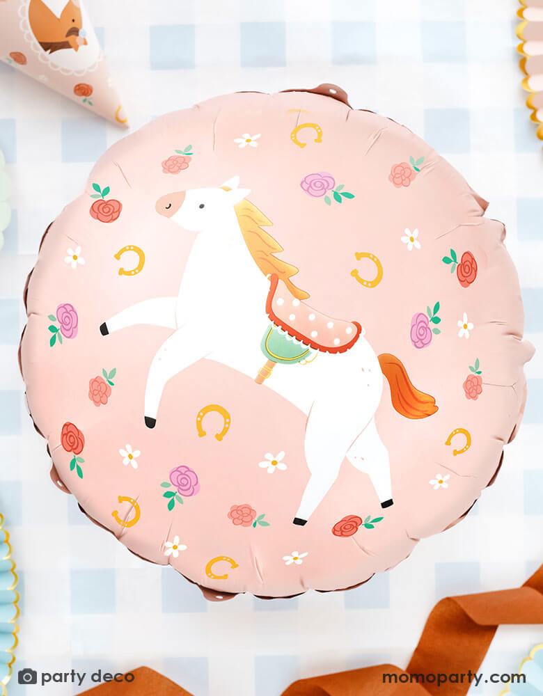 Momo Party's 18" Junior Floral Pony Foil Balloon by Party Deco on a party table with blue gingham tablecloth with party hats and ribbons around it. In an adorable pink, this junior sized round balloon with floral and pony designs is perfect for girl's horse themed birthday parties.