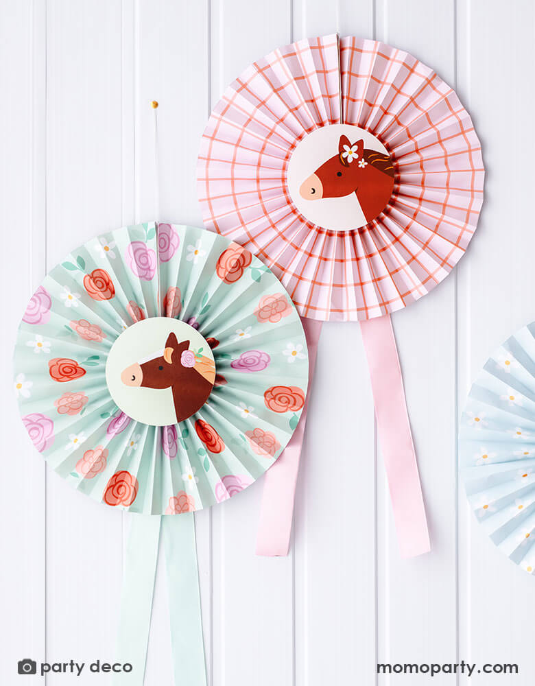 Momo Party's Floral Pony Party Paper Fans by Party Deco hung on a white wall. Comes in a set of 3 in 3 different colors of pink, blue and green and adorable pony design, these party fans feature charming floral designs and adorable pastel ponies. Perfect for adding a playful touch to any celebration, these fans make for a unique and fun decoration.