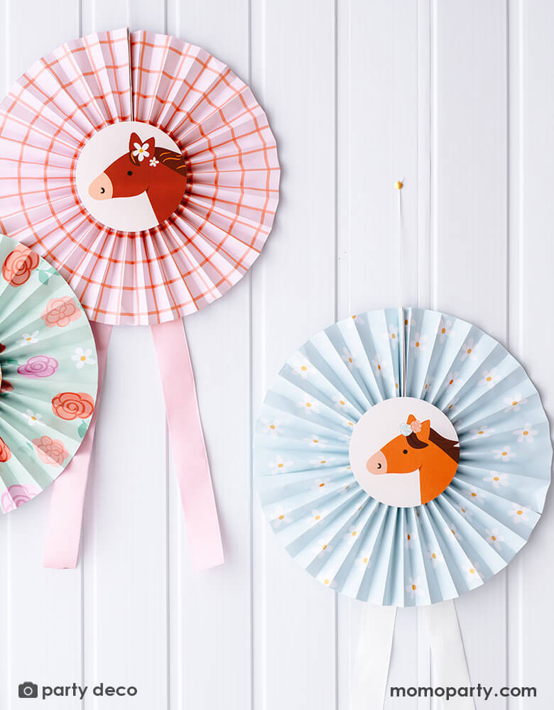 Momo Party's Floral Pony Party Paper Fans by Party Deco hung on a white wall. Comes in a set of 3 in 3 different colors of pink, blue and green and adorable pony design, these party fans feature charming floral designs and adorable pastel ponies. Perfect for adding a playful touch to any celebration, these fans make for a unique and fun decoration.