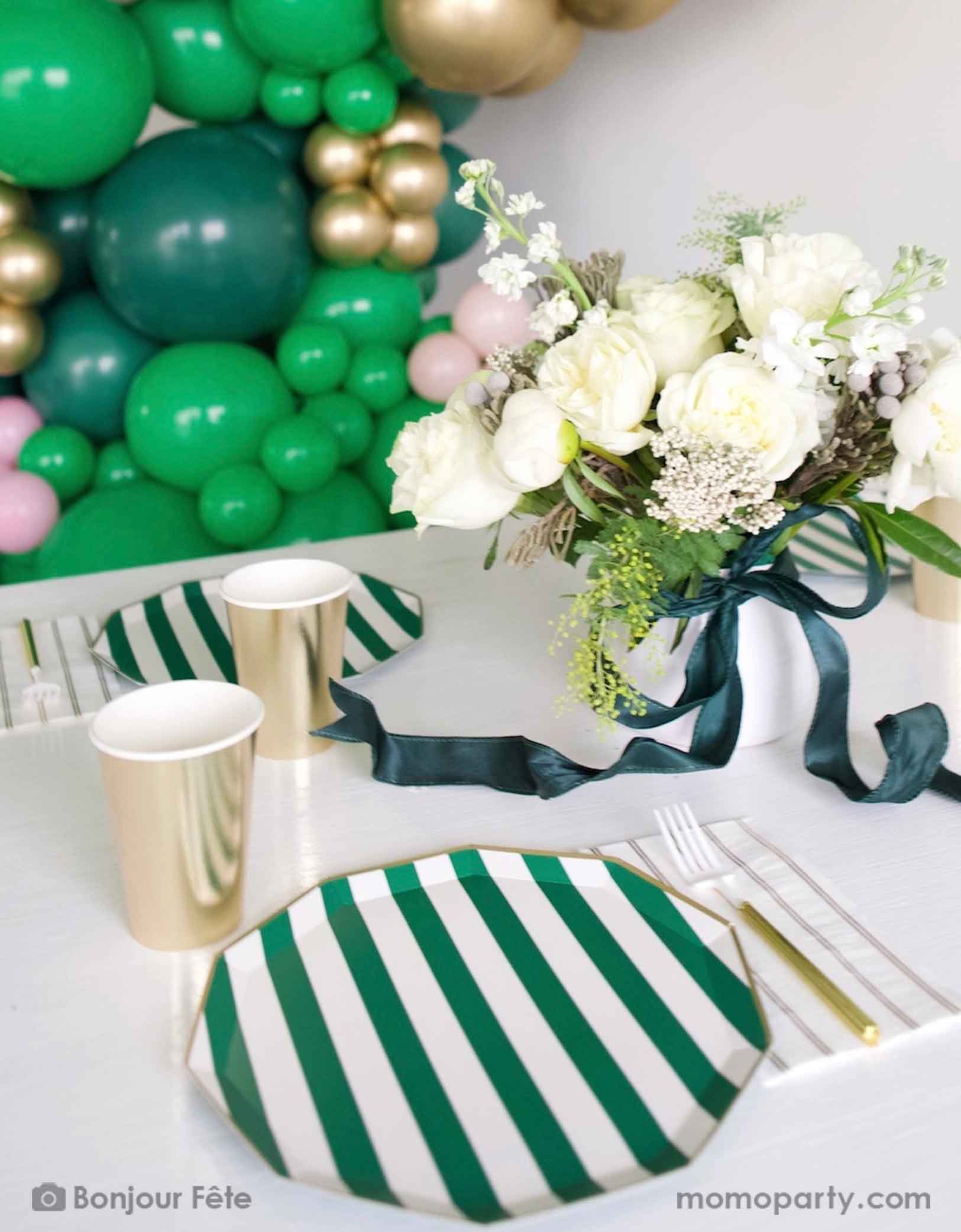 An elegant football themed party table featuring Momo Party's emerald green and cabana stripe and gold tableware including plates, party cups, napkins and gold utensils. With an elegant flower bouquet in white and green with a dark green satin ribbon bow on the vase, along with a balloon wall that consists of green, pink and gold balloons, this is a party inspiration for a girly football, Super Bowl, or a sports themed celebration. 