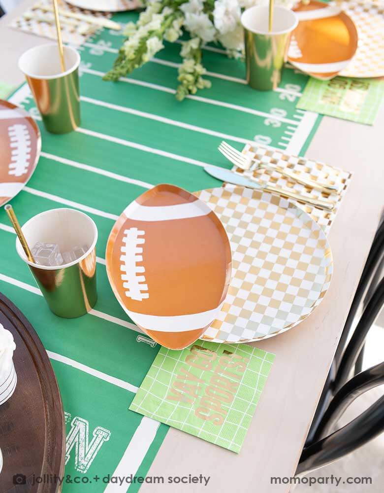 A modern football party table filled with Momo Party's football themed party tableware by Daydream Society including the die-cut football shaped plate, gold and white checkered dinner plate with green and coordinating napkins and gold party cups. In the center of the table there's a football field table runner and a beautiful flower bouquet, all makes great inspirations for football themed celebrations, a Super Bowl watching party or a Sunday football viewing party. 