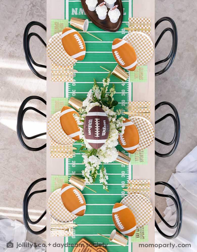 A overhead shot of a long dining table featuring Momo Party's football themed party supplies, including the die-cut football shaped plate, gold and white checkered dinner plate with green and coordinating napkins and gold party cups. In the center of the table there's a football field table runner and a beautiful flower bouquet, all makes great inspirations for football themed birthday celebrations or a Super Bowl, tailgate party.