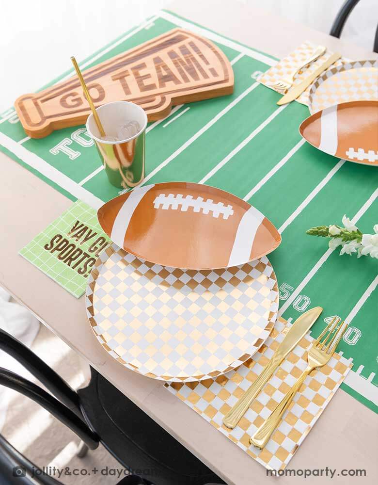 A modern football party table filled with Momo Party's football themed party tableware by Daydream Society including the die-cut football shaped plate, gold and white checkered dinner plate with green napkins says YAY GO SPORTS and gold party cups. In the center of the table there's a football field table runner and a megaphone shaped snack board says GO TEAM! Making this a great inspirations for football themed celebrations, a Super Bowl watching party or a Sunday football viewing party. 