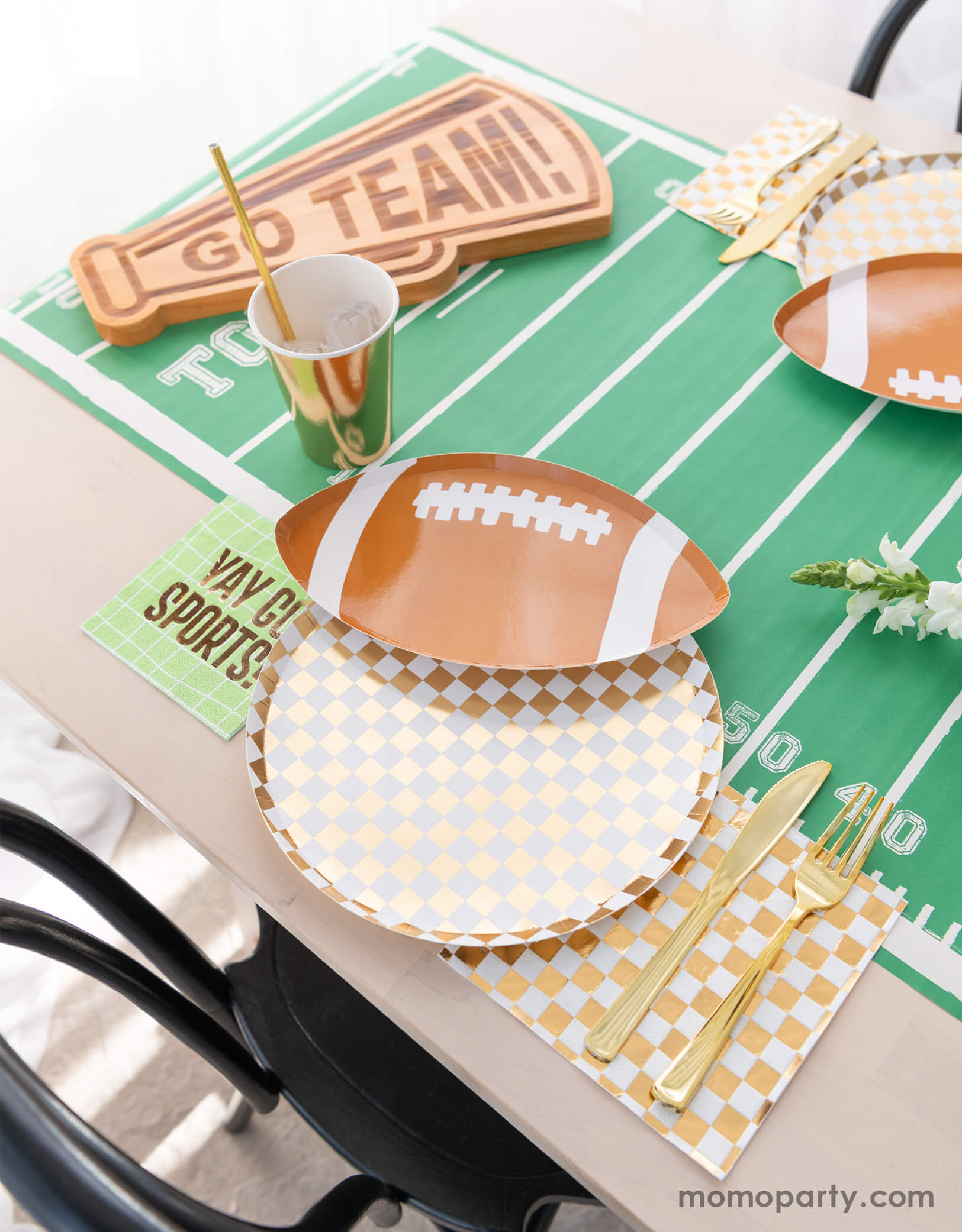 A modern football party table filled with Momo Party's football themed party tableware by Daydream Society including the die-cut football shaped plate, gold and white checkered dinner plate with green napkins says YAY GO SPORTS and gold party cups. In the center of the table there's a football field table runner and a megaphone shaped snack board says GO TEAM! Making this a great inspirations for football themed celebrations, a Super Bowl watching party or a Sunday football viewing party. 