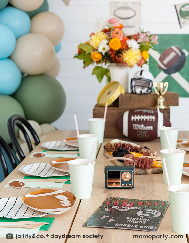 A modern kid's football birthday party table filled with Momo Party's football themed party tableware by Daydream Society including the die-cut football shaped plates, black striped plates, green and mint napkins and party cups. In the center of the table there's a cheese charcuterie board and some decorations like a football, a trophy and a football helmet as the centerpiece. On the wall there are some old school football posters and color matching balloons to add some festivities to the celebration!