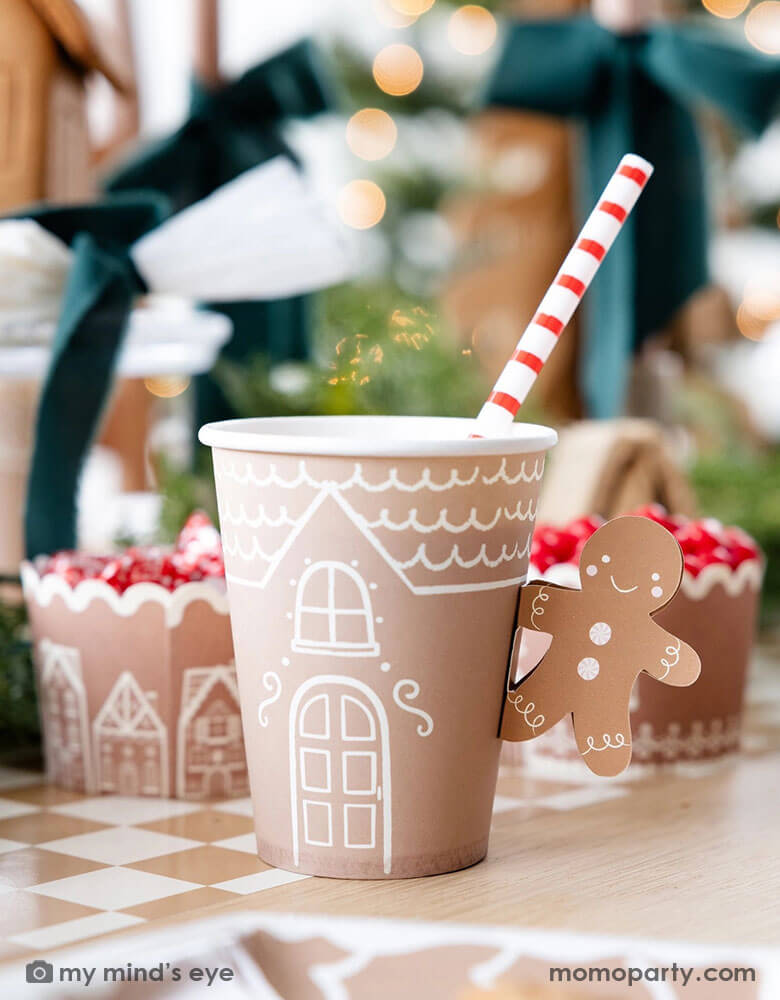 A festive gingerbread themed Holiday party table set up featuring gingerbread themed party tableware by Momo Party including the 12 oz Gingerbread Handled Paper Cup with a festive gingerbread man handle, gingerbread house treat cups filled with peppermints, the mocha checkered napkins and paper table runner. With gingerbread houses and Christmas trees in the back this creates a cozy yet festive vibe for a whimsical Holiday party for kids.