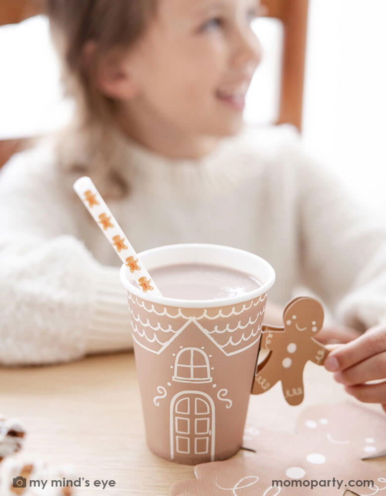 A girl in a white sweater holding Momo Party's 12 oz Gingerbread Handled Paper Cup with a festive gingerbread man handle in a gingerbread house themed Holiday party. With gingerbread man shaped napkins on the table this creates a cozy yet festive vibe for a whimsical Holiday party for kids.