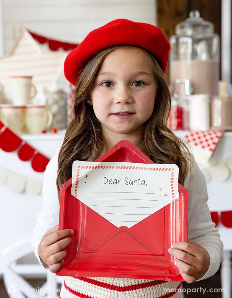 A school aged girl dressed in her Holiday outfit with a red beret holding Momo Party's Letter to Santa envelope shaped plate in front of a hot chocolate station of a table filled with Momo Party's Dear Santa party supplies including a red felt party garland, Santa shaped party cups, red checkered party plates and napkins next to hot cocoa and toppings, making this a cozy yet whimsical set up for a festive Holiday celebration!