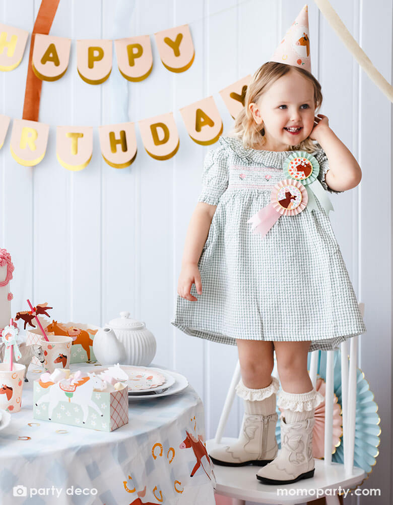 A preschool girl with a birthday party hat in a gingham dress and Momo Party's horse rosette ribbons on her chest, standing next to a party table featuring Momo Party's floral pony themed party supplies including floral pony party cups, plates, cupcake toppers, napkins treat boxes and paper straws, along with the blush happy birthday party banner hung on the wall - making this a perfect inspo for a girl's horse themed birthday party.