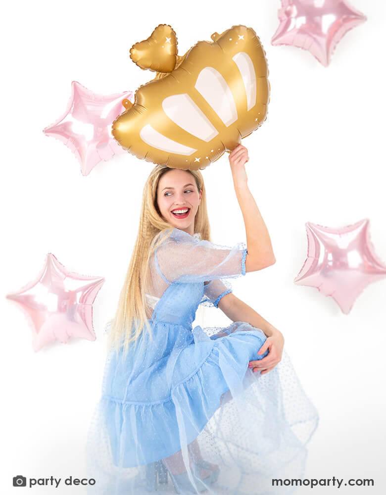 A blonde lady in a Cinderella-like light blue tutu dress holding Momo Party's 20" x 17" princess crown shaped foil balloon by Party Deco. In the back there are four pink star shaped foil balloons as the background, making this a good inspiration for a princess themed party decoration. 