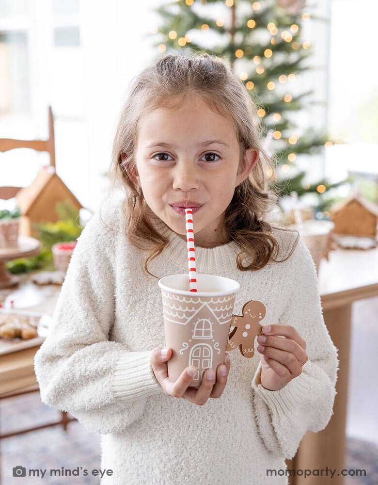 A cute girl in a white sweater holding drinking hot chocolate from Momo Party's 12 oz Gingerbread Handled Paper Cup with a festive gingerbread man handle.  She's sipping her drinks with a red and white striped reusable straw. In her back there's a table filled with gingerbread houses and a Christmas tree with lights in background, which creates a cozy yet festive vibe for a whimsical Christmas party for kids this Holiday season.