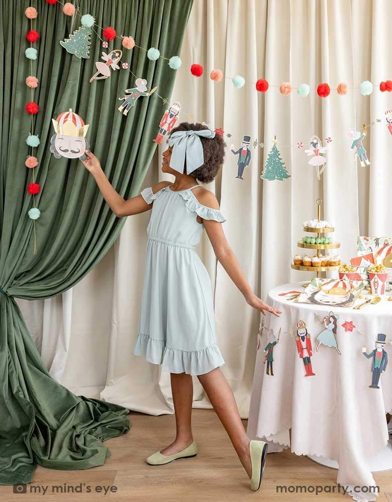 A school aged girl doing a ballet dance pose holding Momo Party's 10" Nutcracker head shaped party plate in the air, standing next to a party table filled with Holiday treats and Nutcracker themed party supplies including plates, cups, napkins and party garland. In her back there's a stage curtain in satin green and beige which is decorated with Momo Party's Nutcracker garland with pom-poms which adds a festive touch to the Holiday decoration.