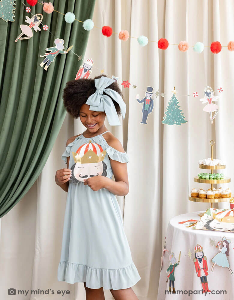 A school aged girl holding Momo Party's 10" Nutcracker head shaped party plate standing next to a party table filled with Holiday treats and Nutcracker themed party supplies. In her back there's a stage curtain in satin green and beige which is decorated with Momo Party's Nutcracker garland with pom-poms which adds a festive touch to the Holiday decoration.