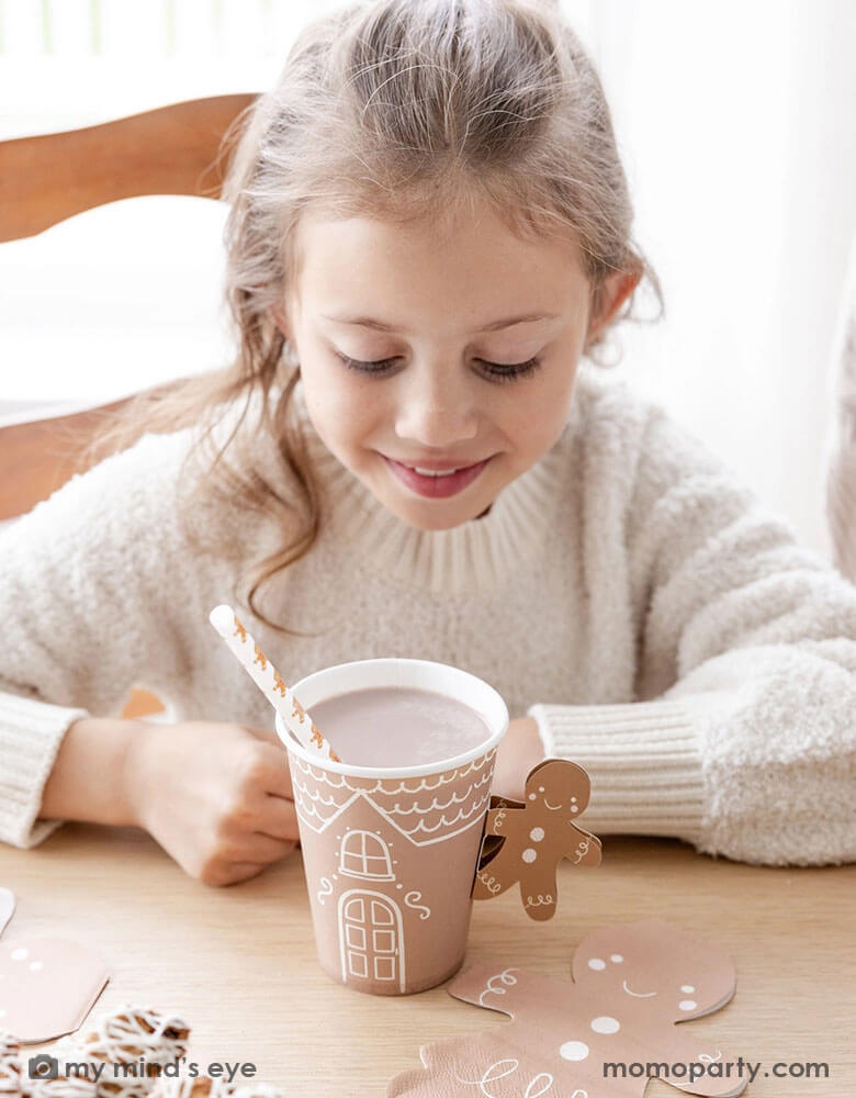 A girl in a white sweater looking into Momo Party's 12 oz Gingerbread Handled Paper Cup with a festive gingerbread man handle. There's hot chocolate in the cup with a gingerbread man straw, in a gingerbread house themed Holiday party. With gingerbread man shaped napkins and Holiday treats on the table this creates a cozy yet festive vibe for a whimsical Holiday party for kids.