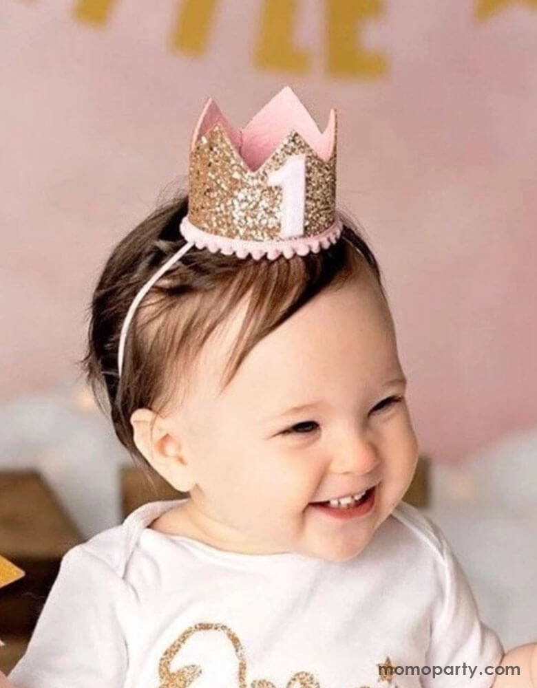 A one-year girl in her pale pink outfits wears Momo Party's 3" first birthday pink glittered birthday party crown. She has a sweet smile and in her back there's a pink party backdrop with gold happy birthday banner in the back.