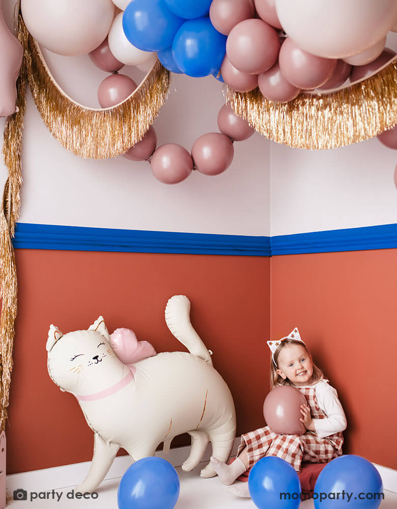 A girl dressed like a cat sitting next to the kitty cat shaped foil balloon from Momo Party. Around them the room is decorated with festive balloons in different shades of pink and royal blue witch matches the colors of the walls. Along with gold tinsel fringe garlands, it creates a fun and festive atmosphere for kid's pet or cat themed birthday celebration.