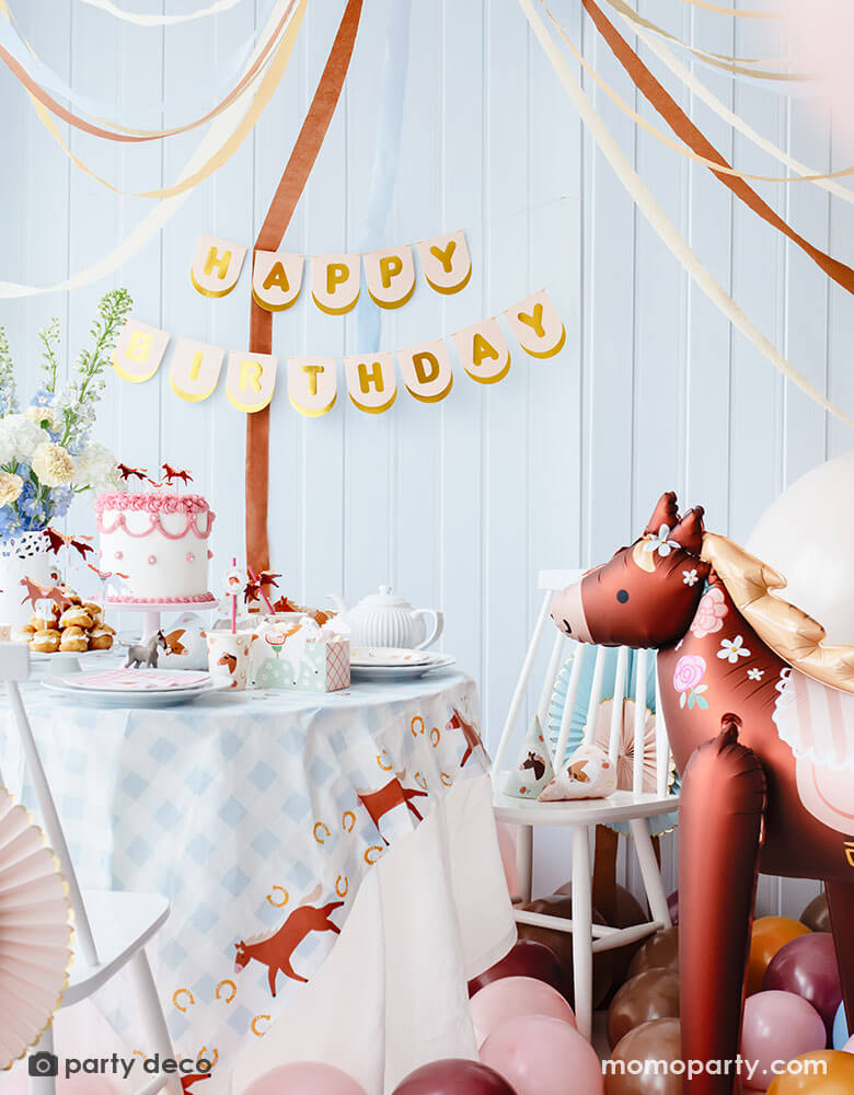 A pony themed birthday party table featuring Momo Party's floral pony party supplies including party cups, plates, cupcake toppers, napkins treat boxes and paper straws. In the middle of the table is a pink and white buttercream cake topped with pony shaped birthday candles, along with the blush happy birthday party banner hung on the wall. Next to the table stands an adorable pony shaped foil balloon adorned with floral designs. Making this a perfect inspo for girl's horse themed birthday celebration.