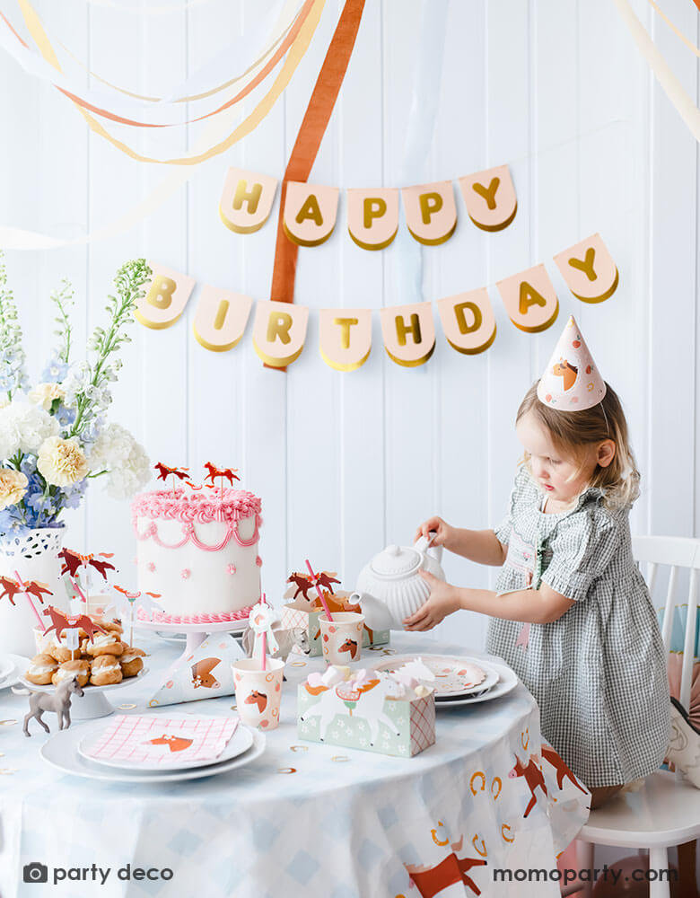 A preschool girl with a birthday party hat in a gingham dress pouring tea from a tea pot on a party table featuring Momo Party's floral pony themed party supplies including floral pony party cups, plates, cupcake toppers, napkins treat boxes and paper straws. In the middle of the table is a pink and white buttercream cake topped with pony shaped birthday candles, along with the blush happy birthday party banner hung on the wall - making this a perfect info for a girl's horse themed birthday party.