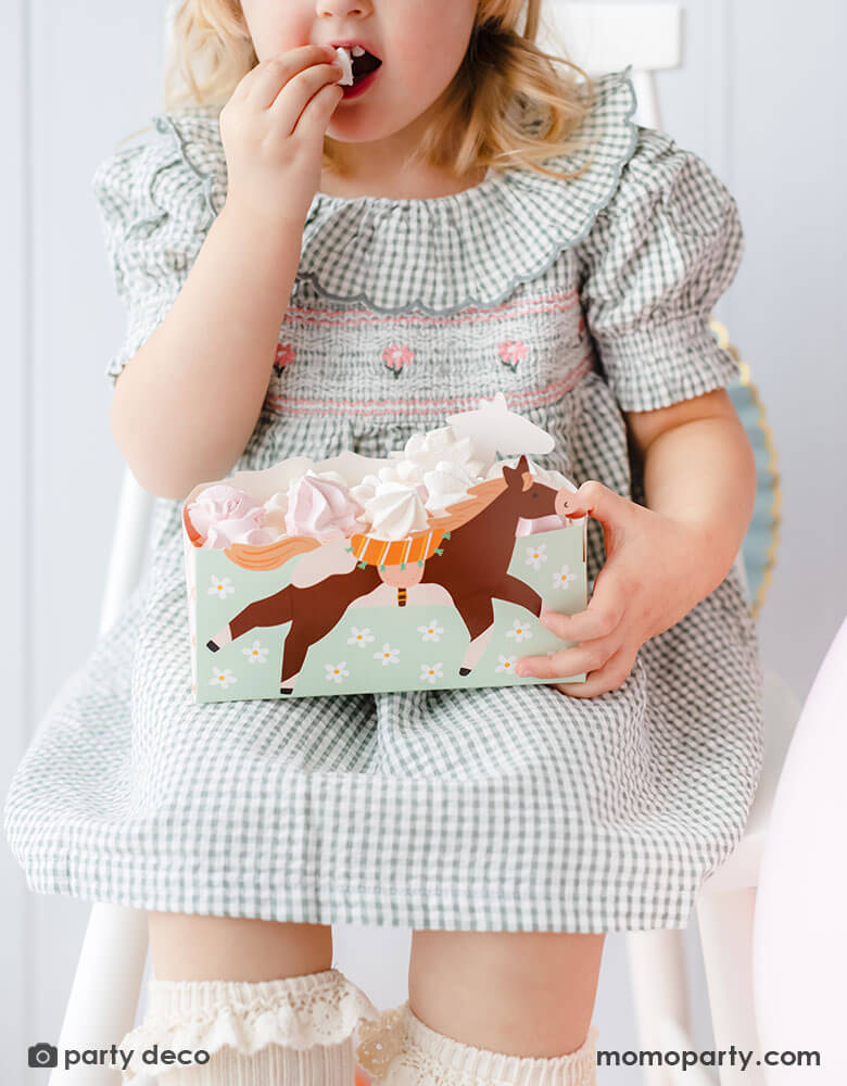 A preschool aged girl in a green gingham checkered dress sitting on a chair holding Momo Party's floral pony treat box by Party Deco. In the snack box are pink and white meringue - a great party snack idea for girl's horse themed birthday party.