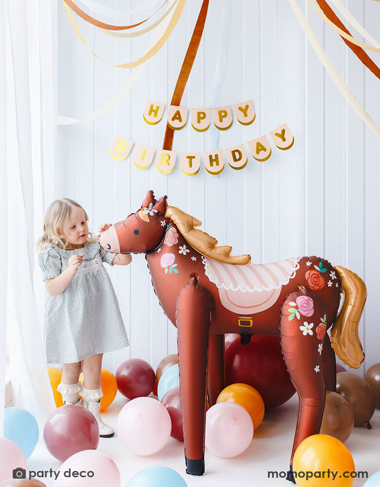 A preschool aged girl petting Momo Party's 40" x 42" standing floral pony foil balloon in a pony themed birthday party room with Happy Birthday paper banner hung on the wall. Around the standing pony foil balloon, there are multiple latex balloons in difference sizes and matching colors lay around to make a festive scene for a birthday celebration.