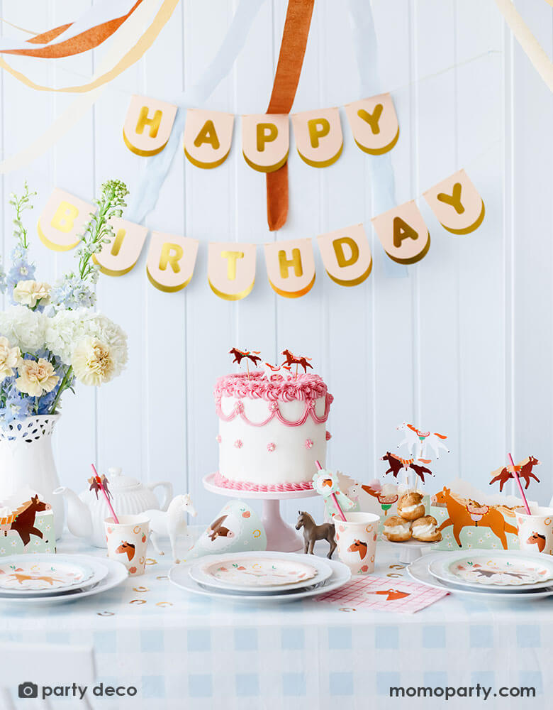 A kid's pony horse themed birthday party table decoration featuring Momo Party's floral pony themed party supplies including floral pony party cups, plates, cupcake toppers, napkins treat boxes and paper straws. In the middle of the table is a pink and white buttercream cake topped with pony shaped birthday candles, along with the blush happy birthday party banner hung on the wall and floral arrangement, this table setting makes a great inspo for a charming horse themed birthday celebration for girls. 