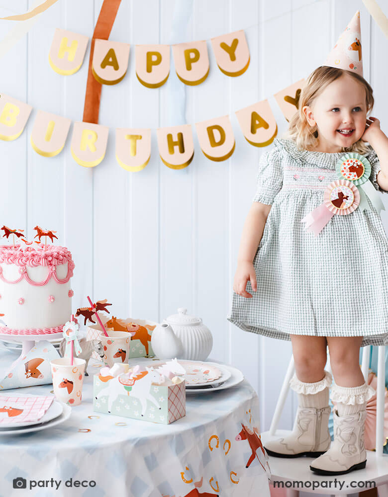 A preschool girl with a birthday party hat in a gingham dress and blush cowgirl boots standing next to a party table featuring Momo Party's floral pony themed party supplies including floral pony party cups, plates, cupcake toppers, napkins treat boxes and paper straws. In the middle of the table is a pink and white buttercream cake topped with pony shaped birthday candles, along with the blush happy birthday party banner hung on the wall - a perfect info for a girl's horse themed birthday party.