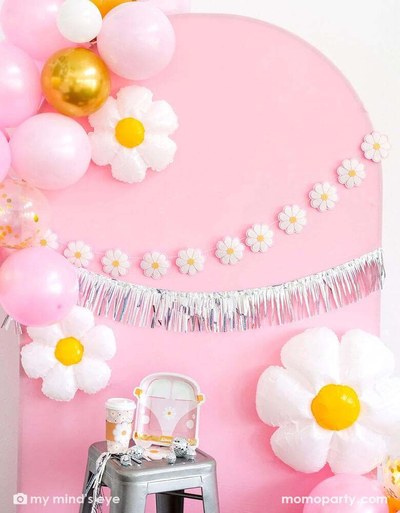 A pink retro daisy themed party decoration set up featuring Momo Party's disco daisy themed party supplies including VW van shaped plates, party cups, daisy shaped napkins on the stool. In the back there's a pink curve wooden backdrop adorned with My Mind's Eye's 6ft Disco Daisy Puffy Felt Banner and a silver fringe garland. Along the backdrop there are also a balloon garland with gold, pink and white balloons and daisy shaped foil balloon. Making this a great inspo for a groovy daisy hippie themed bash!