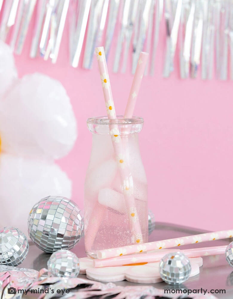 A milk bottle with pink soda and Momo Party's disco daisy reusable straws. Around the bottle there are Momo Party's disco daisy napkins and silver disco ball decorations. Above the the tableware is silver tinsel fringe garland, perfect for a groovy retro celebration!